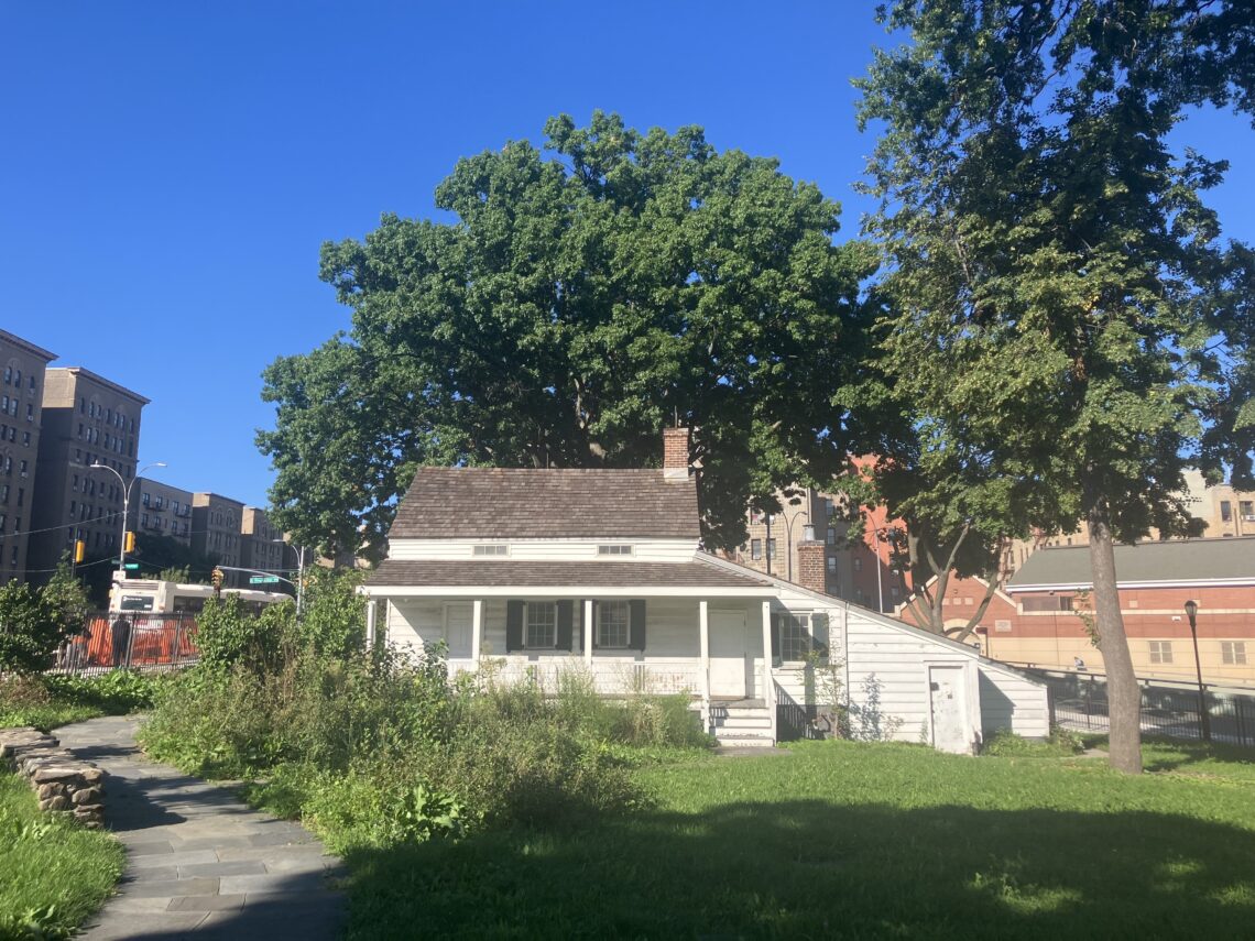 Un piccolo cottage di legno bianco con il portico, circondato da alberi e prati, con i palazzi del Bronx sullo sfondo