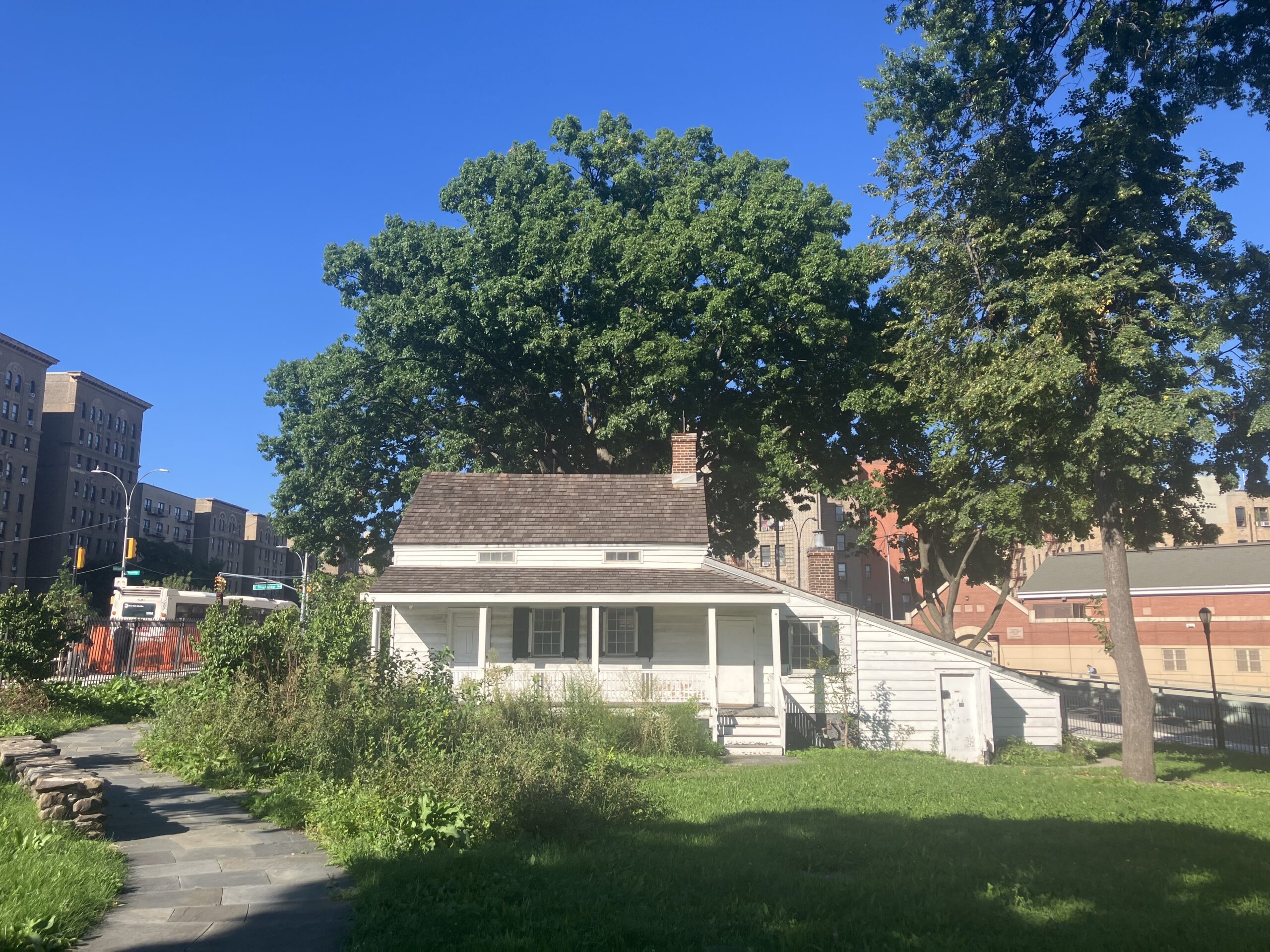 Un piccolo cottage di legno bianco con il portico, circondato da alberi e prati, con i palazzi del Bronx sullo sfondo
