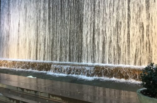 Un piccolo parco urbano lastricato con sedie e tavolini bianchi, alberi che fanno ombra e, sul fondo, una parete attraversata da una cascata d'acqua continua, incastonato tra due palazzi di Manhattan