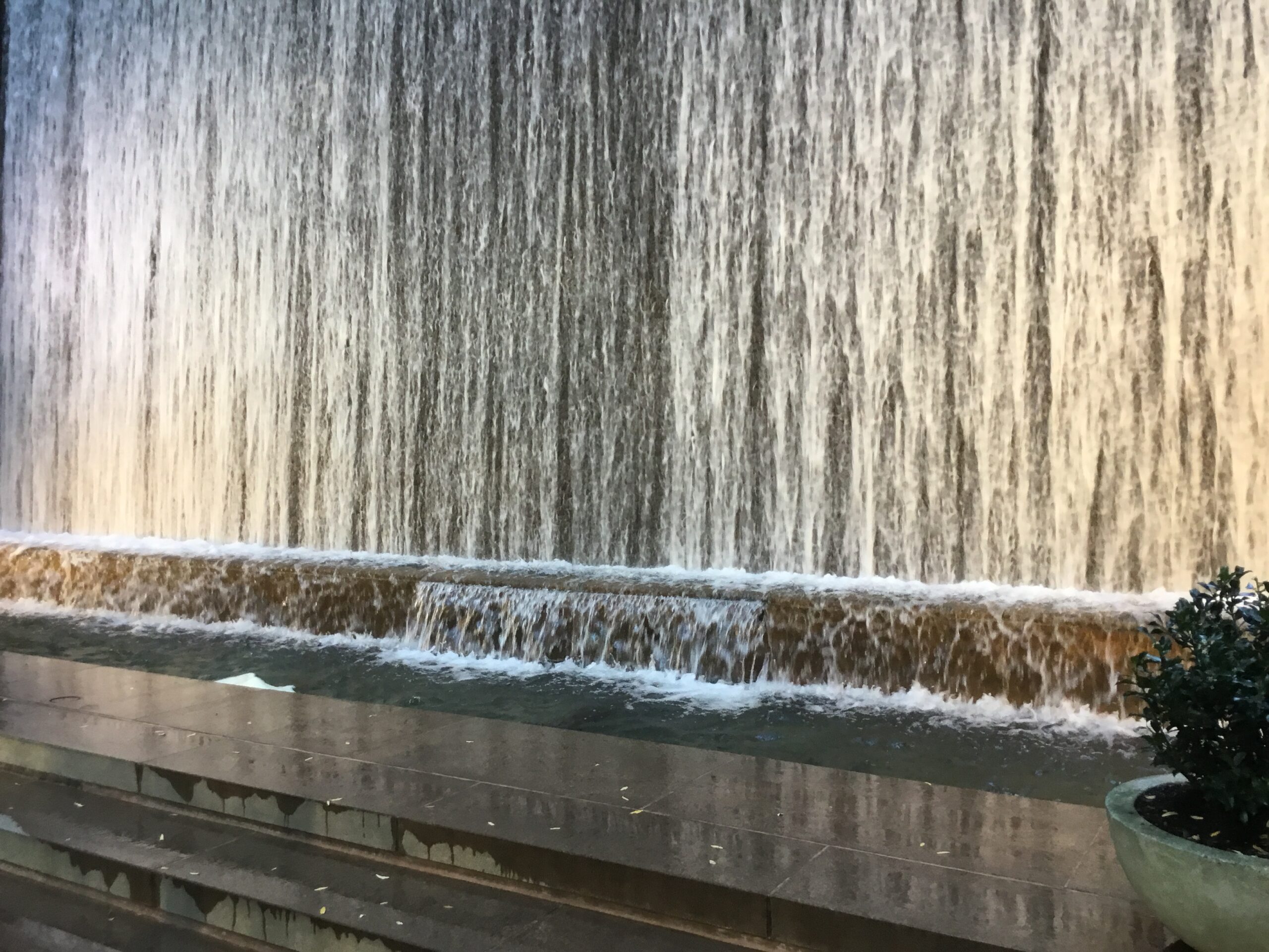 Un piccolo parco urbano lastricato con sedie e tavolini bianchi, alberi che fanno ombra e, sul fondo, una parete attraversata da una cascata d'acqua continua, incastonato tra due palazzi di Manhattan