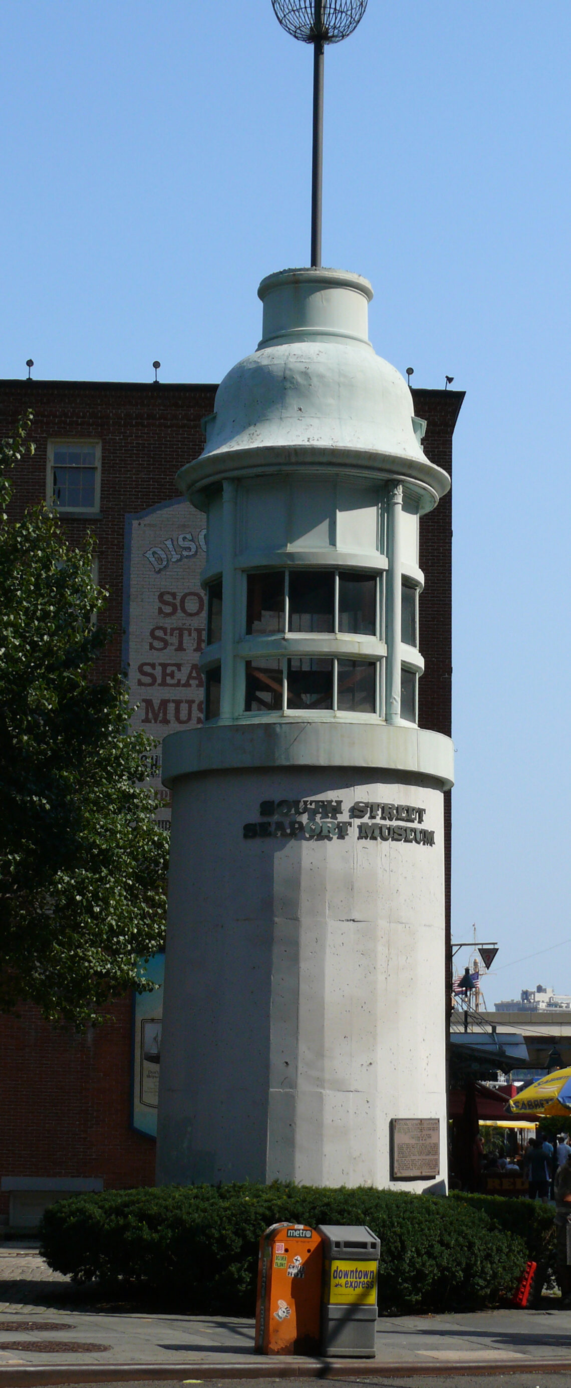 Un piccolo faro conico bianco sormontato da un gabbiotto di vetro e una sfera nera in cima, collocato tra gli edifici del South Street Seaport di New York